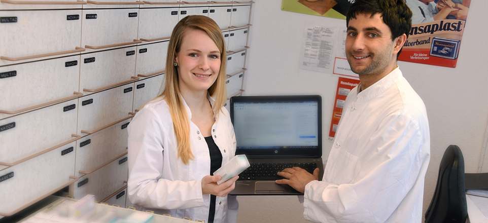 Zwei Mitarbeitende in einer Apotheke mit Laptop und vor Medikamentenregalen