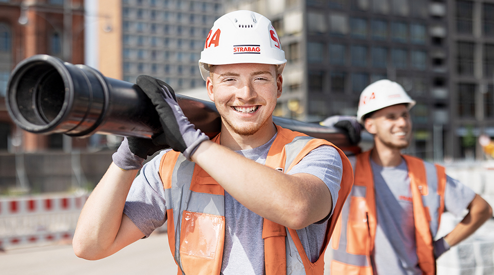 Zwei junge Männer in Arbeitskleidung tragen Rohr