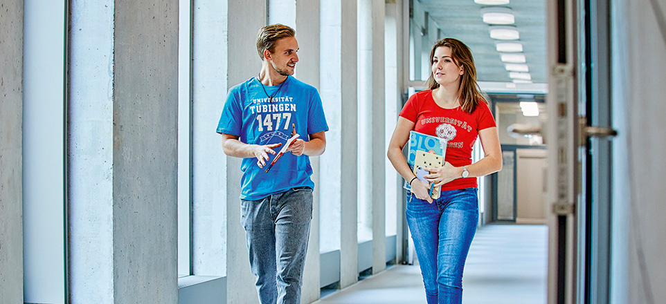 Zwei junge Menschen in T-Shirts der Universität Tübingen gehen lächelnd durch einen hellen Flur und unterhalten sich.
