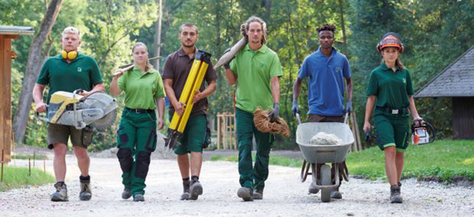 6 Menschen in grüner Arbeitskelidung laufen mit Schubkarre und Gartenwegzeug auf einem Weg in Richtung der Kamera