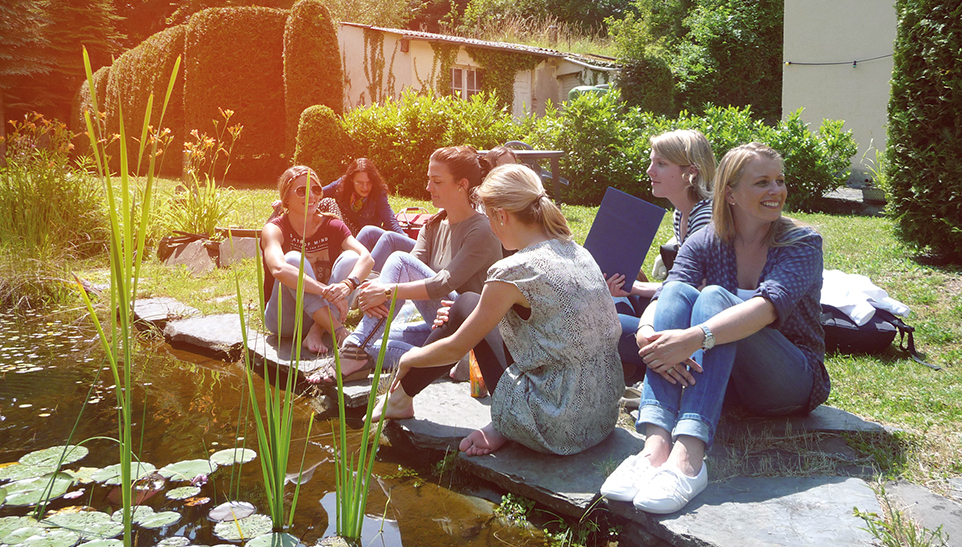 Studierende sitzen enspannt bei sonnigem Wetter an einem kleinen Teich