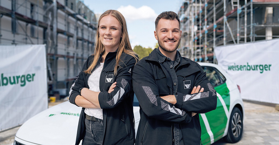 Zwei Personen lächeln in die Kamera und stehen Rücken an Rücken. 