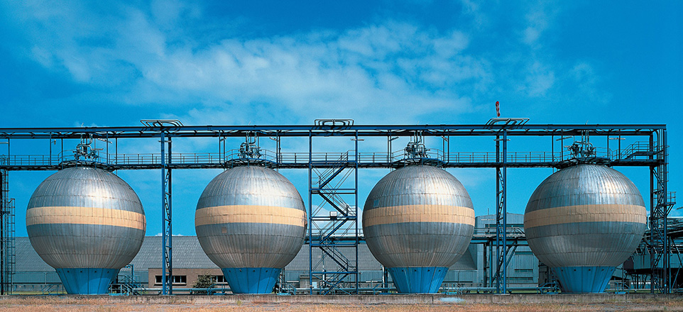 Das Bild zeigt vier große, kugelförmige Industriebehälter (Kugelspeicher), die nebeneinander auf einem Betriebsgelände stehen. Die silberfarbenen Behälter stehen auf blauen Trägerkonstruktionen. 